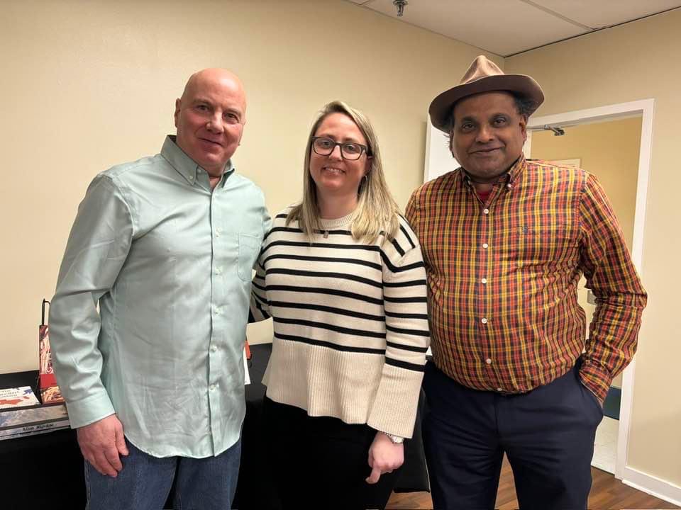 white, bald man in blue button up shirt, white woman with blond hair in white and black striped sweater, and Sri Lankan man in brown and white plaid button-up shirt with a tan hat